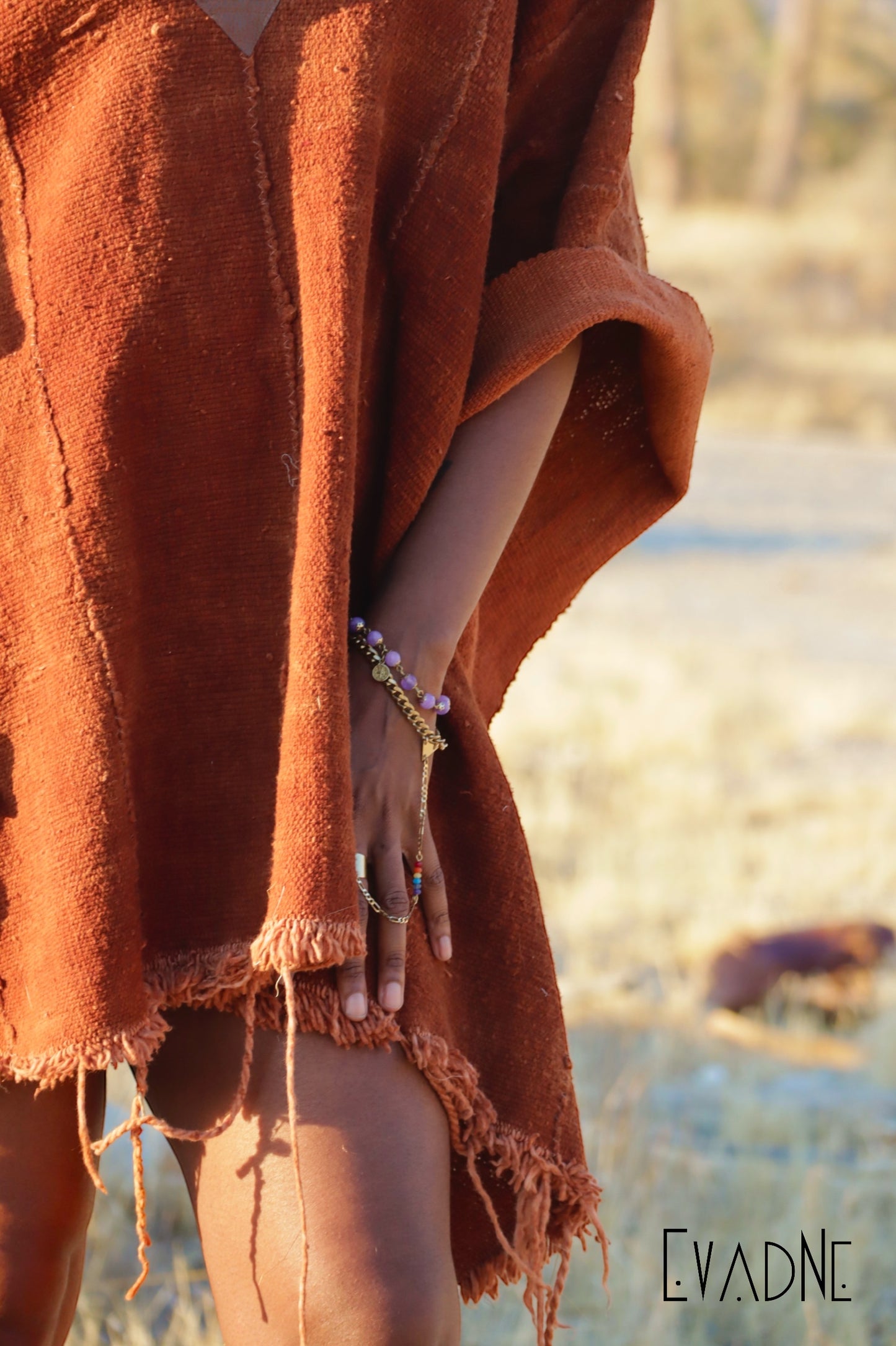 Person wrapped in a rust colored poncho with bracelets on and a blurred natural background