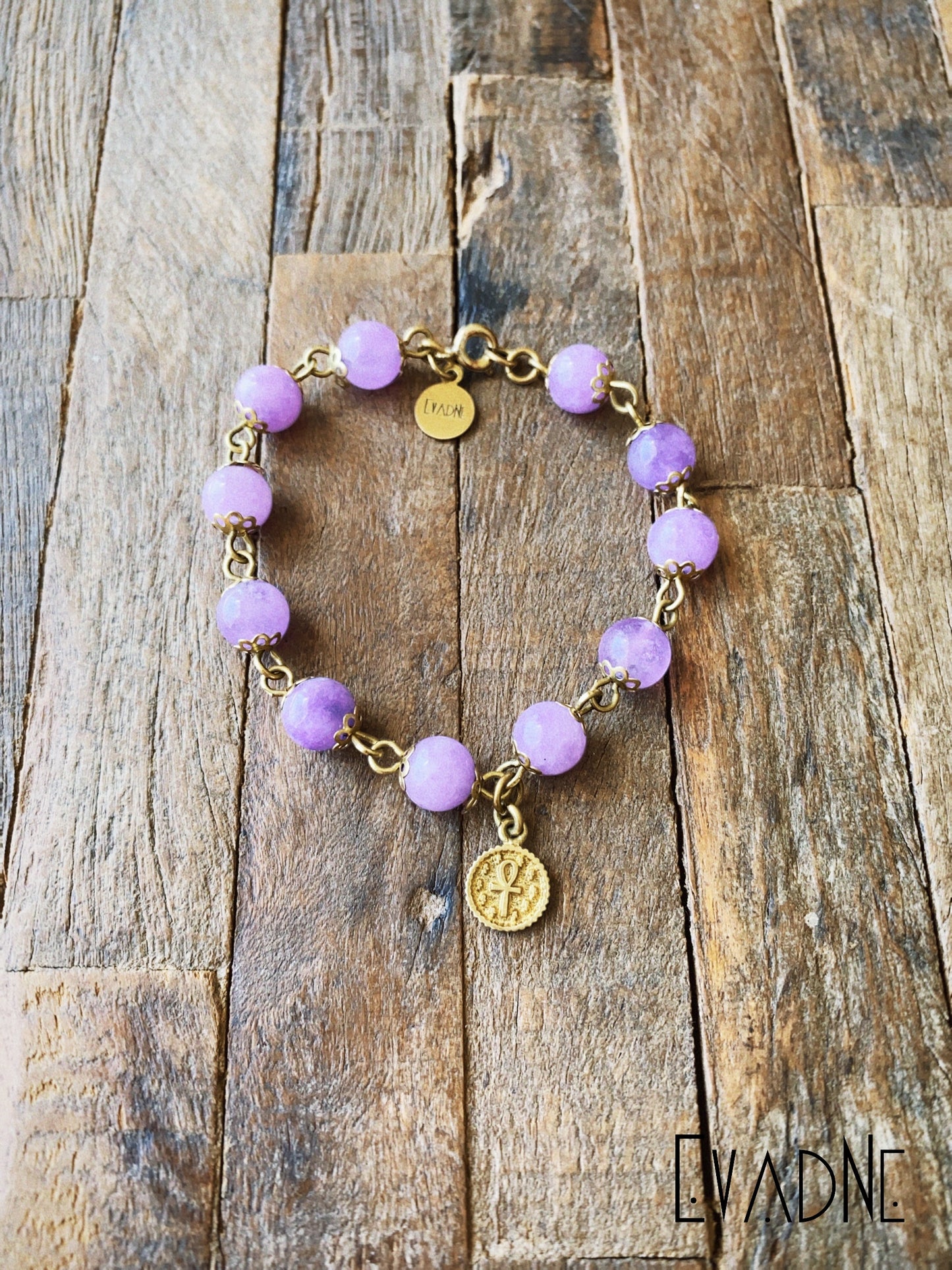 Amethyst beaded bracelet with gold chain and charms on a wooden surface, branded 'Evadne'.