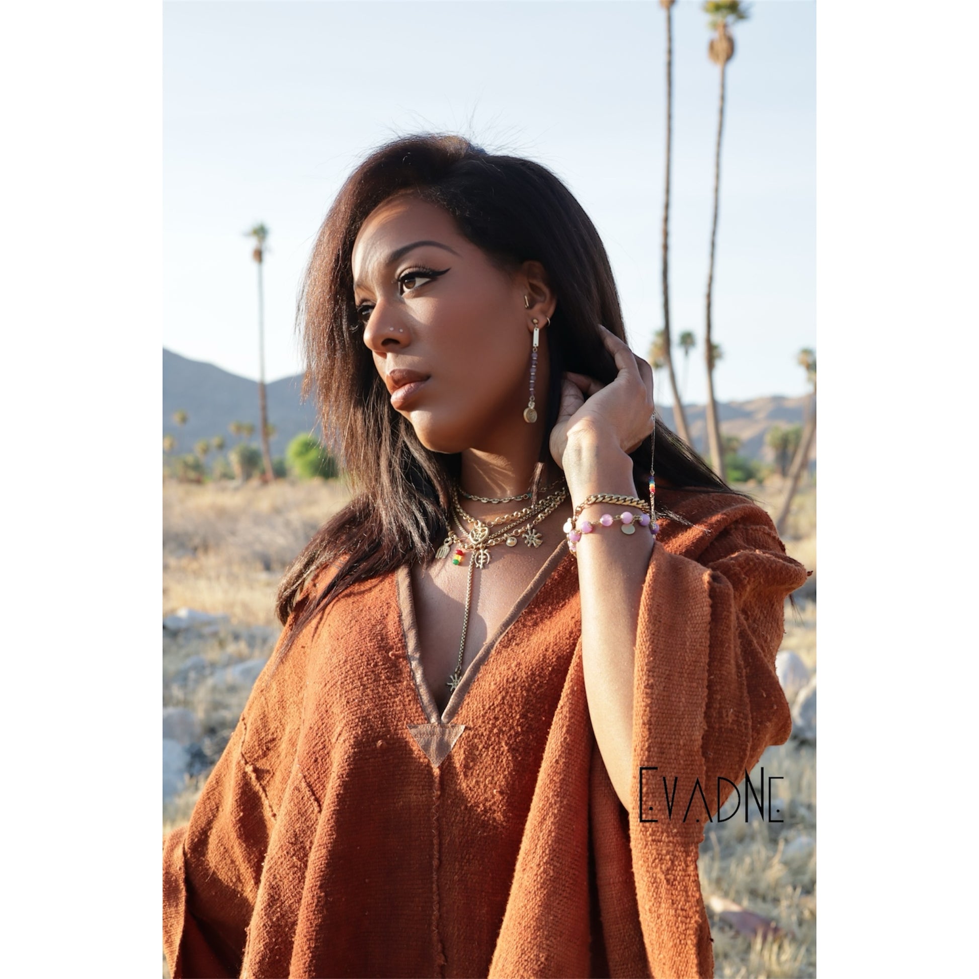 Woman in a desert setting with palm trees, wearing a brown outfit and minimalist amethyst gemstone earrings.