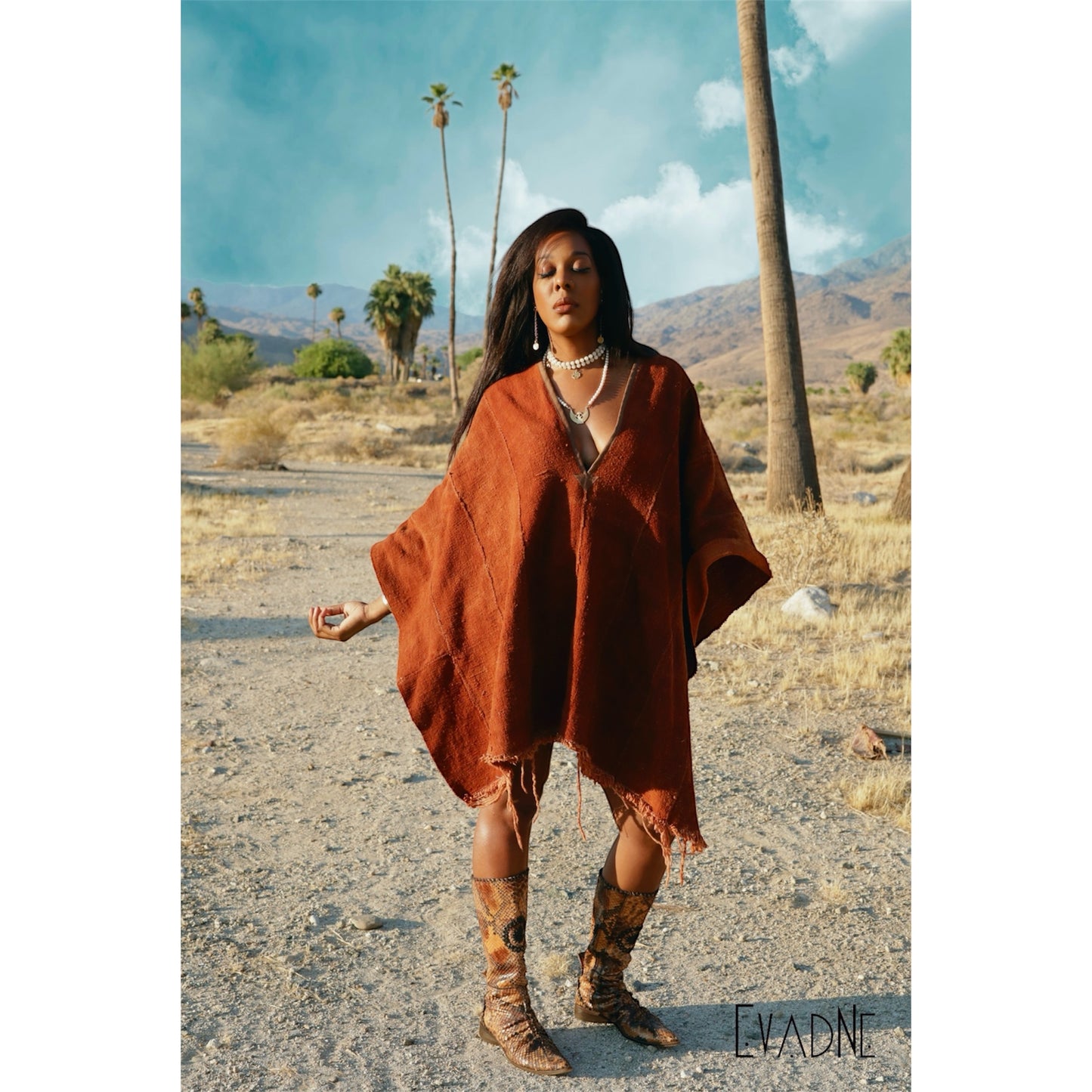 Woman in a brown poncho and Nyame Dua Adinkra charm on white jade protection necklace standing in a desert with palm trees and mountains in the background.
