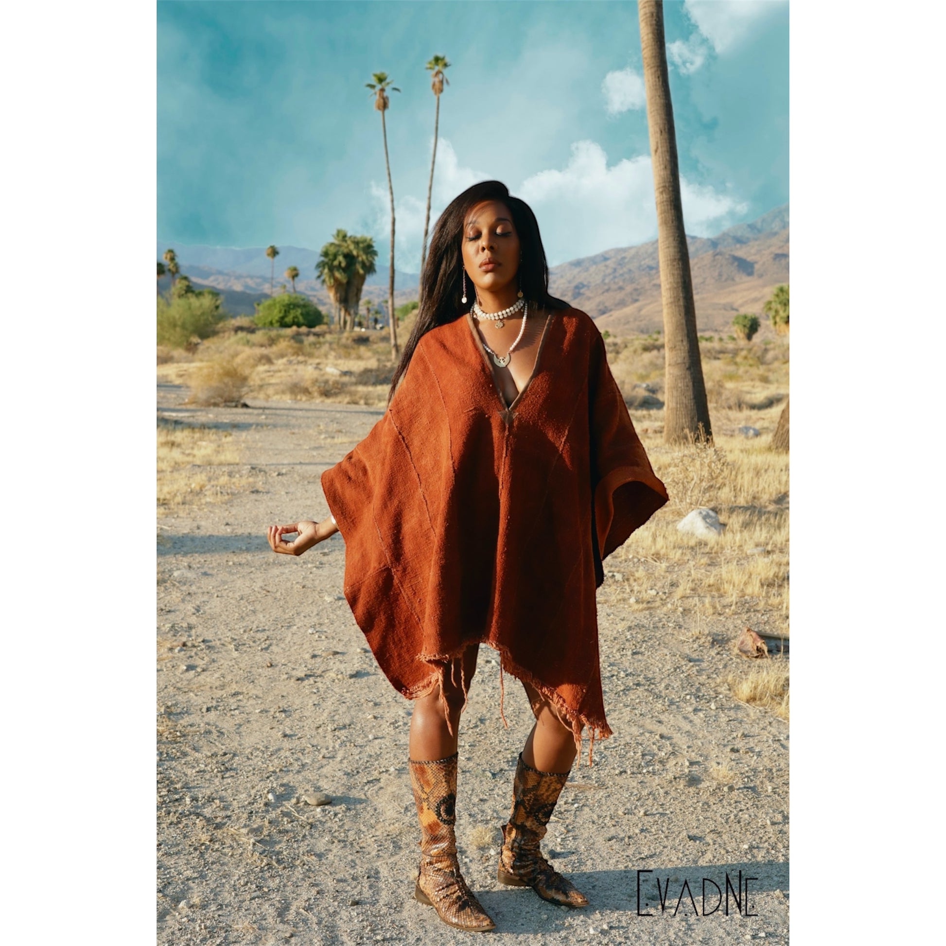 Woman in a brown poncho and Nyame Dua Adinkra charm on white jade protection necklace standing in a desert with palm trees and mountains in the background.